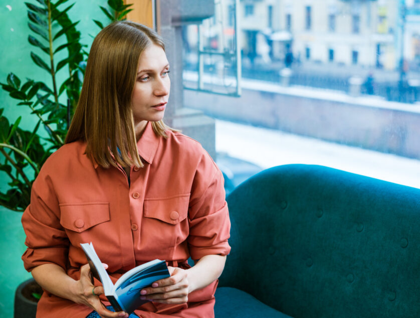 Beautiful caucasian girl spends her free time at home, sitting on couch, Beautiful caucasian girl spends her free time at home, sitting on couch, reading a book and looking thoughtfully out the window. Caucasian woman lifestyle, leisure at home